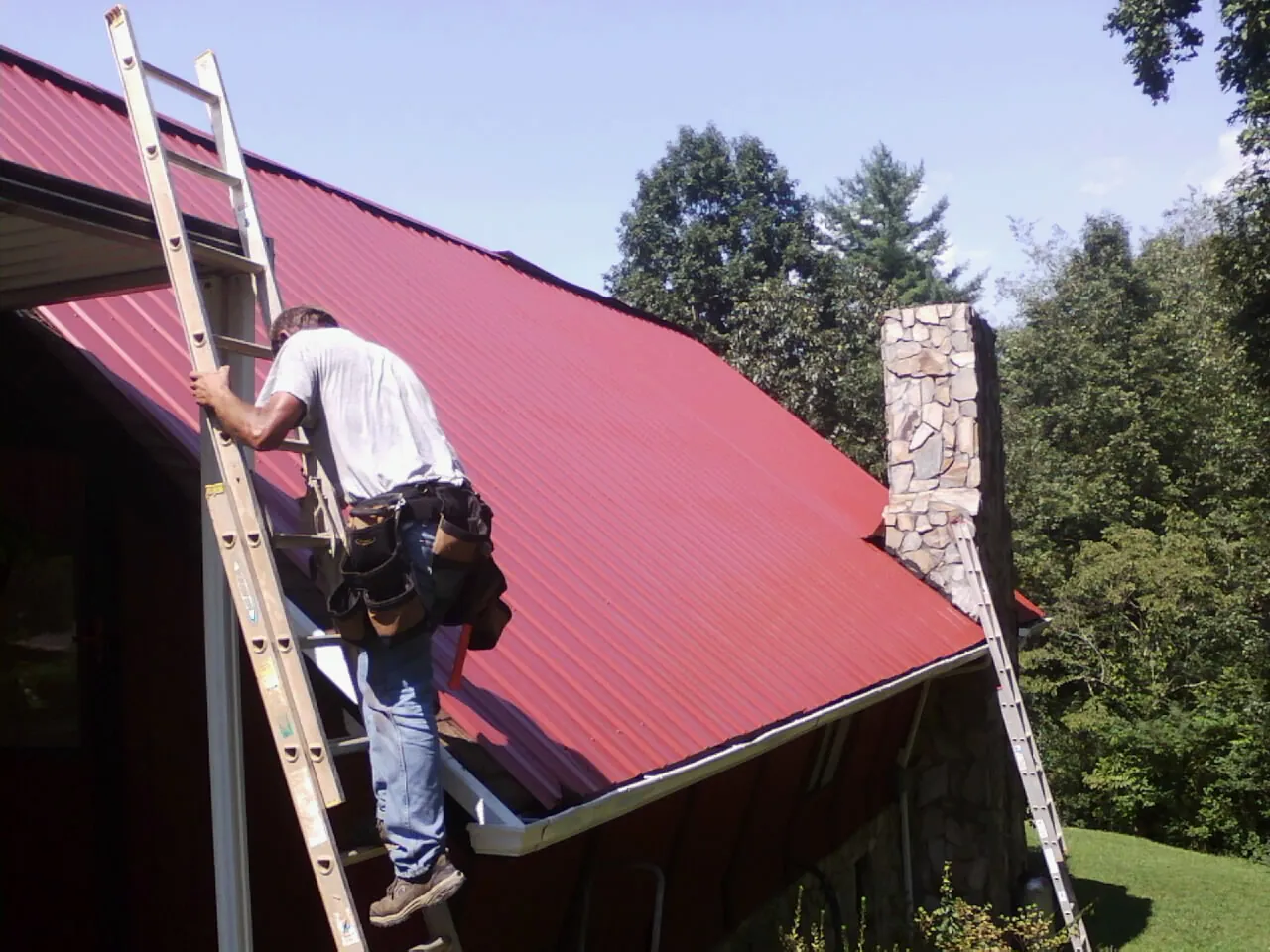 Roofer on a ladder beside a red metal roof and stone chimney — Soffit & Fascia Repair by Roofing Brothers in Windsor